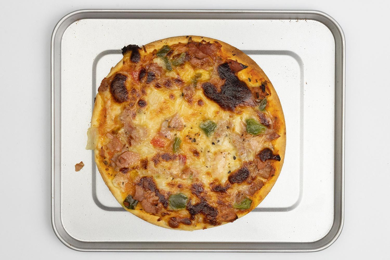 A toaster oven baked 9-inch thick-crust meat pizza with cheese, onions, and green bell peppers on top inside a baking pan.