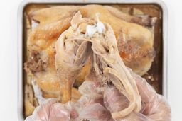 Two hands pulling apart a roasted chicken thigh. In the white background is the rest of the roasted chicken on a baking pan.