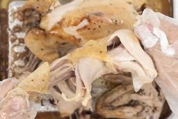 Two hands pulling apart a roasted chicken thigh. In the white background is the rest of the roasted chicken on a baking pan.