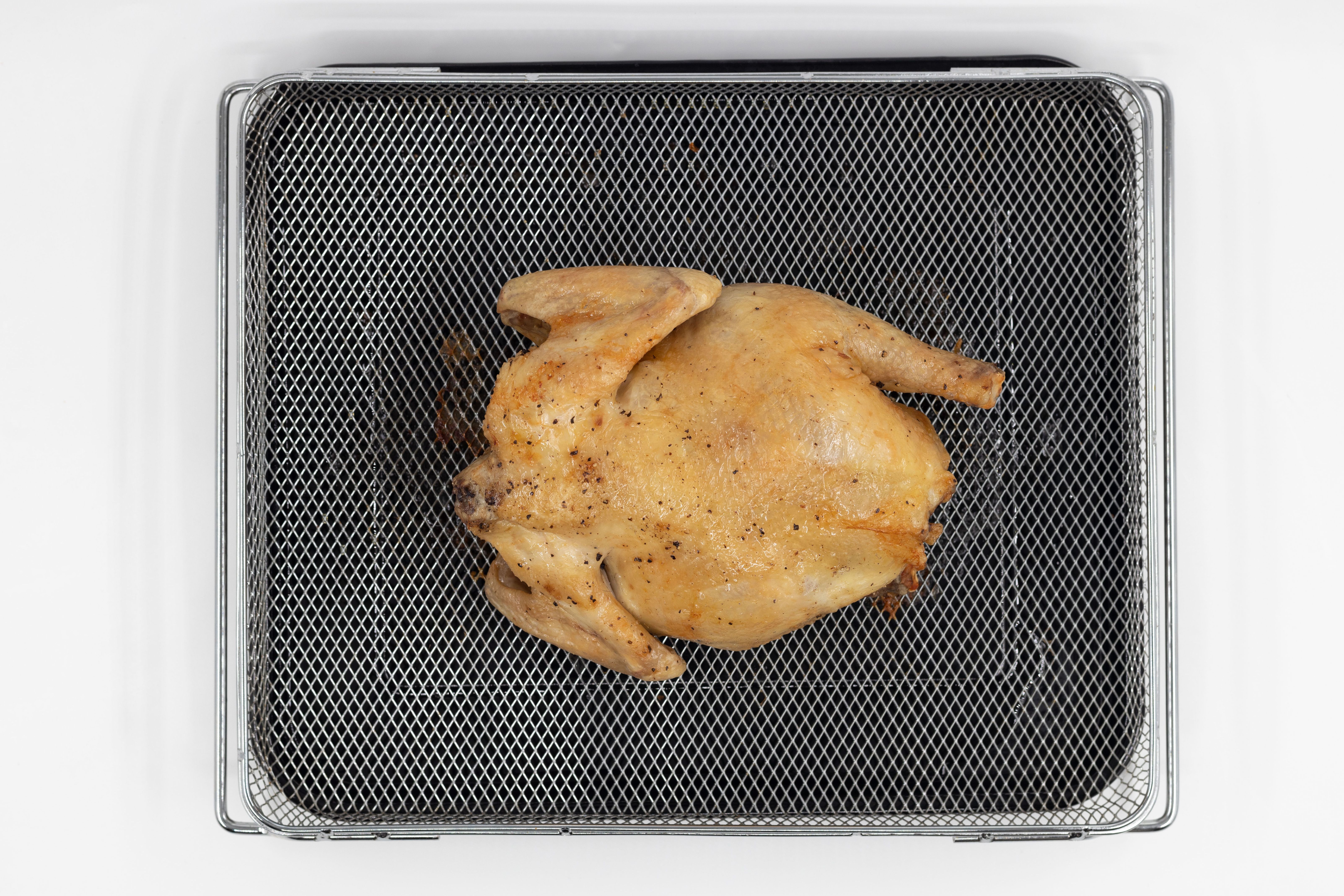 A whole roasted chicken using a toaster oven on a white background. The chicken is backside up inside an air fryer basket on top of a black baking pan.