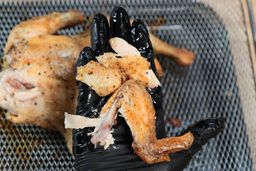 A palm holding a broken up chicken wing showing the cooked meat and golden skin. In the background is the rest of the whole roasted chicken inside an air-fryer basket on top of a baking pan.