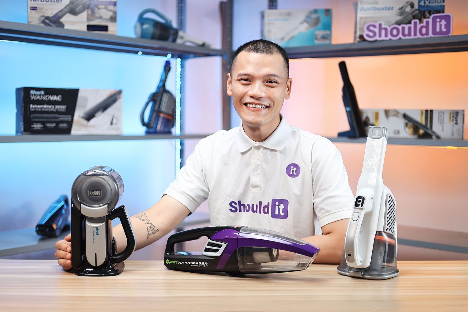 ShouldIT’s reviewer posing with a number of handheld vacuum models. There are some models in the background with a visible ShouldIT logo.