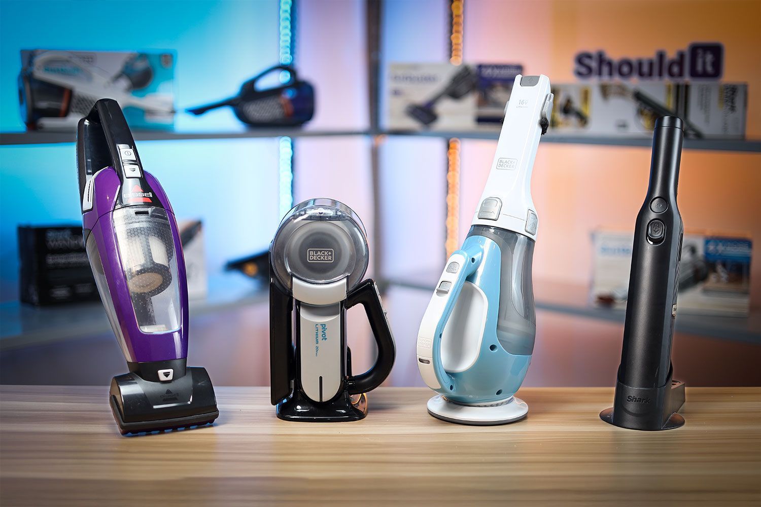 Four different handheld vacuums posed next to each other on a table. Different models are posed in the background with a visible ShouldIT logo