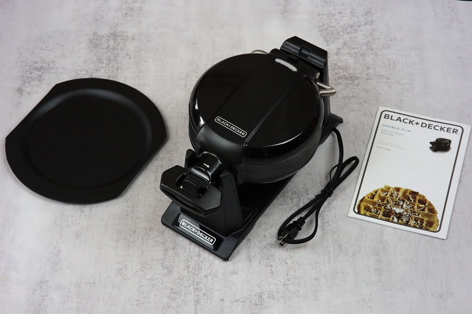 The all-black Black and Decker double waffle maker WMD200B next to its removable drip tray and instruction manual.