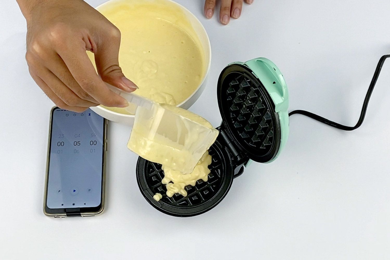 Fresh waffle batter is poured onto the plate of the DASH Mini waffle maker, with a phone displaying a timer to the right.