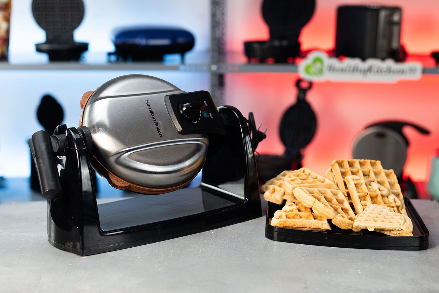 Hamilton Beach 26031 waffle maker with its drip tray filled with freshly-baked waffles against a blurry blue and red background.