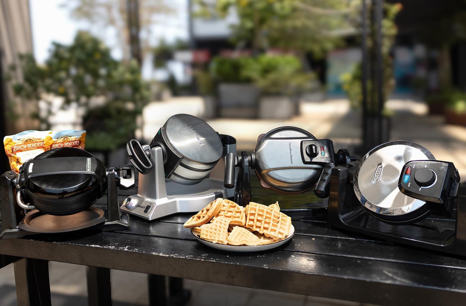 The best Belgian waffle makers on a wooden table against a blurry background, with a plate of fresh plate of waffles in the center.