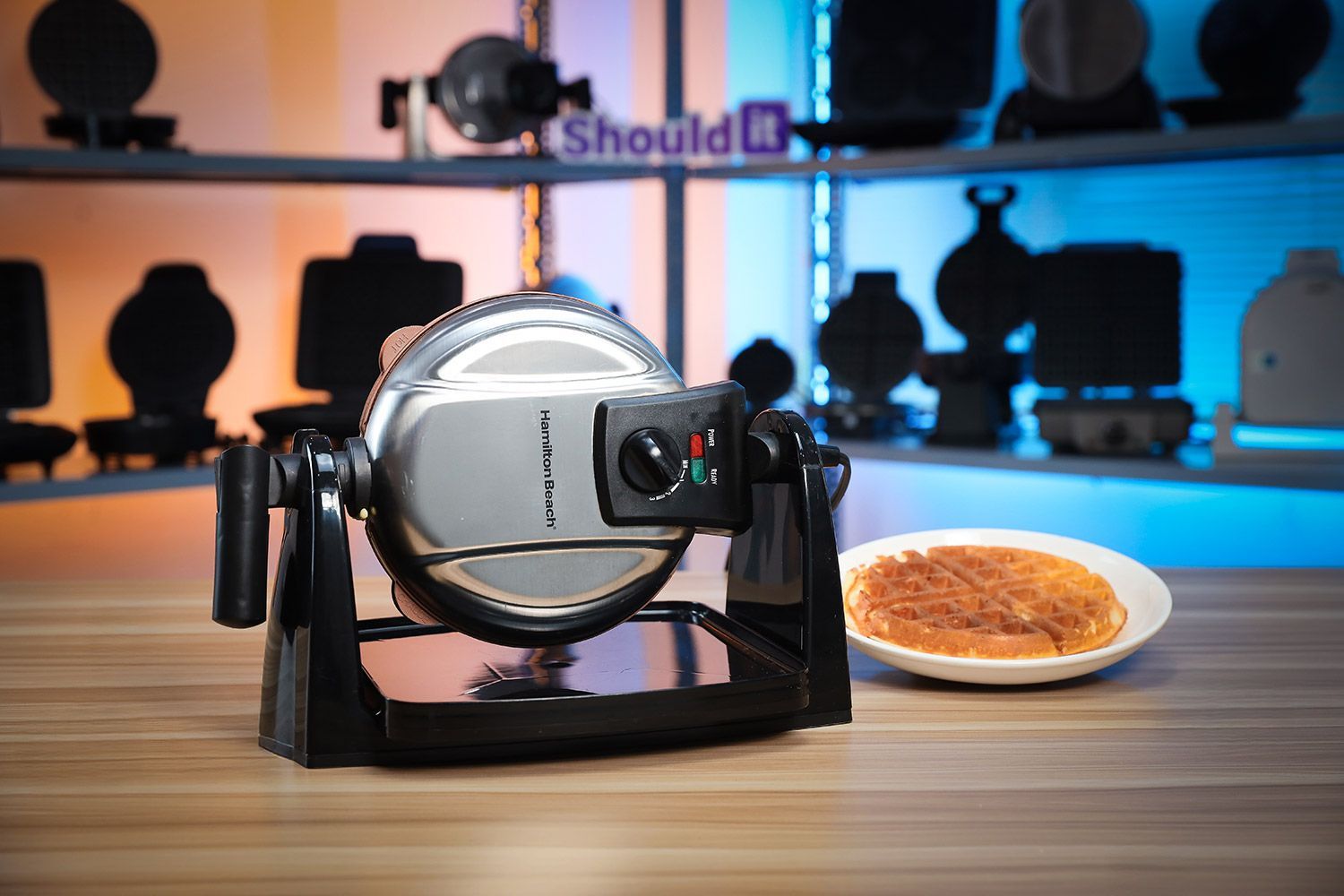 Hamilton Beach 26031 waffle maker with its drip tray filled with freshly-baked waffles against a blurry blue and red background.