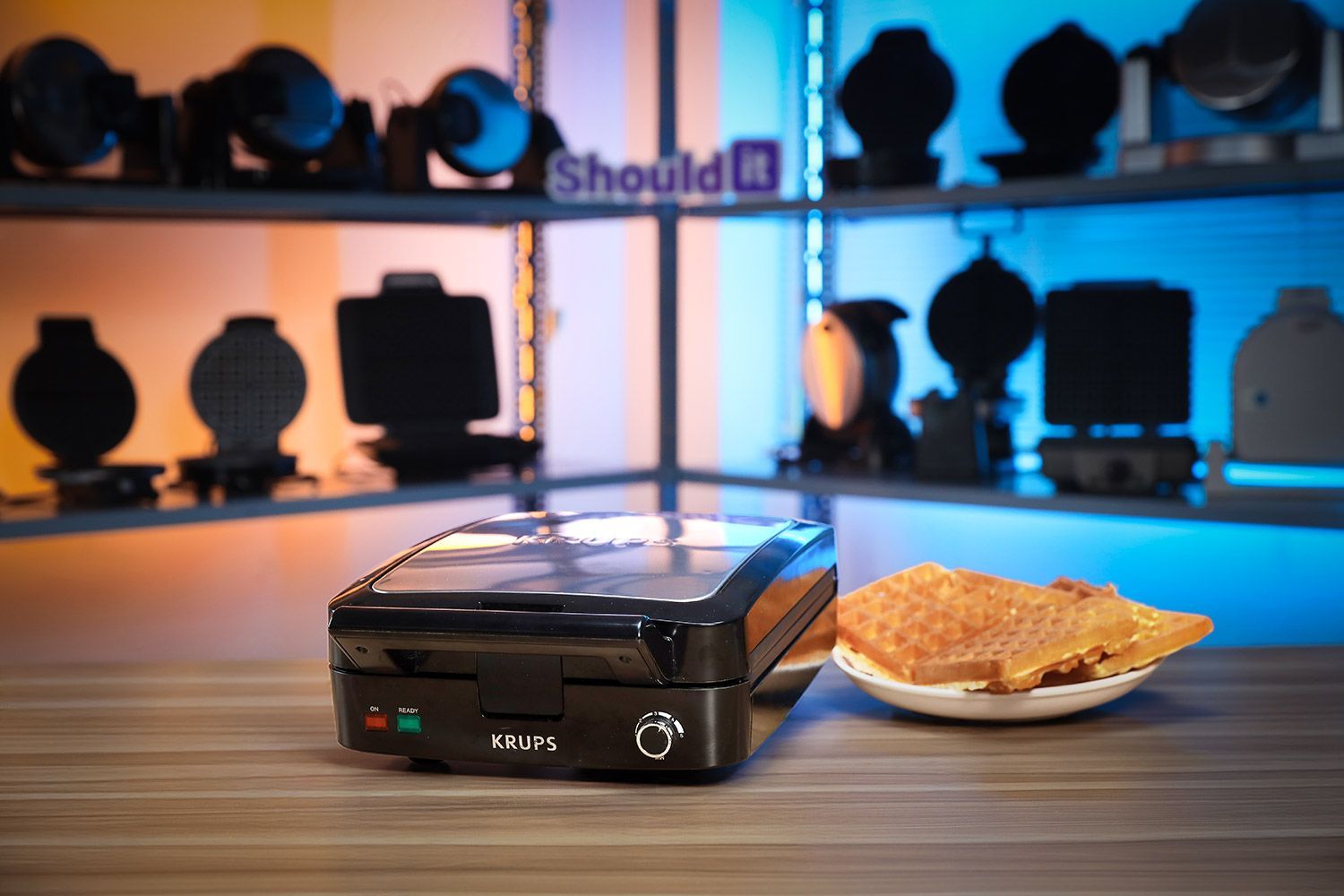 The KRUPS Belgian waffle maker with its lid opened and two waffles on the plate next to a plate of cooked waffles on a wooden countertop