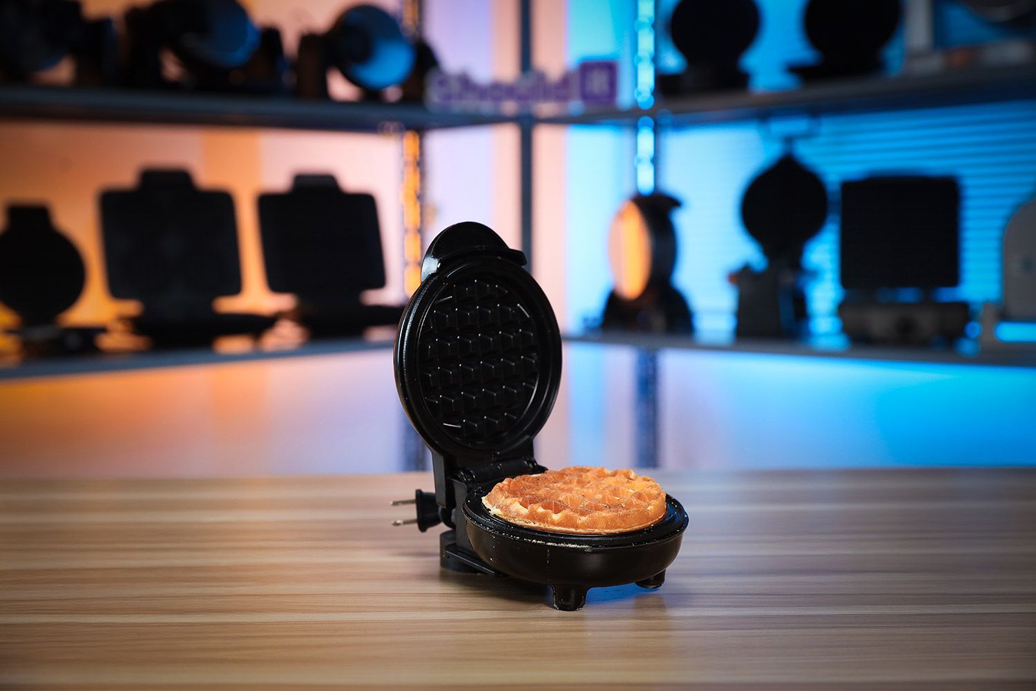 The Holstein Housewares Mini waffle maker next to a plate of cooked mini and 4-inch waffles on a wooden tabletop.