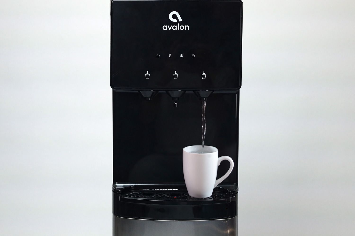 A paper cup filled with water resting on a drip tray below the cold water outlet on a water cooler dispenser.