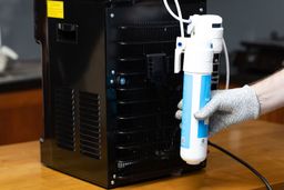 A Gloved hand holding a water filter removed from its bracket at the rear of a bottleless countertop water cooler dispenser.