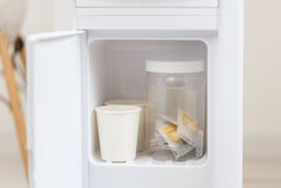 A small storage cabinet on a water cooler dispenser with two paper cups and plastic jar demonstrating the size.