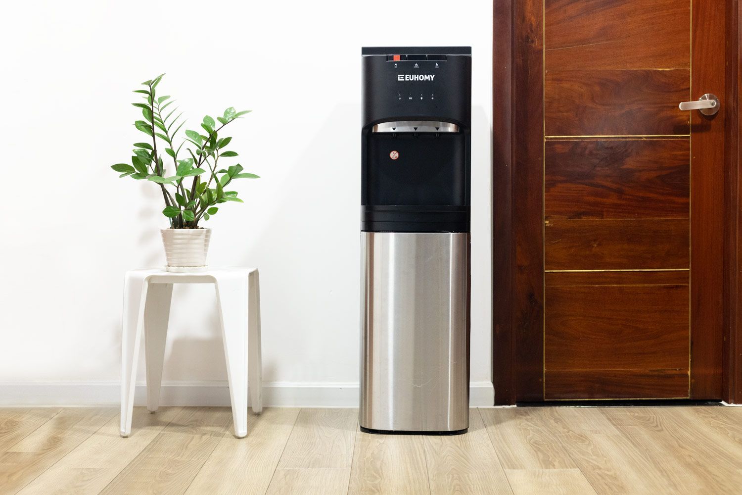 Euhomy bottom loading water cooler dispenser standing next to an office door with a potted plant to the left.