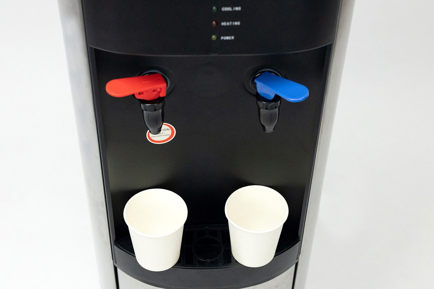 Two small paper cups barely fitting completely on the drip tray of a water cooler dispenser with a faucet design.