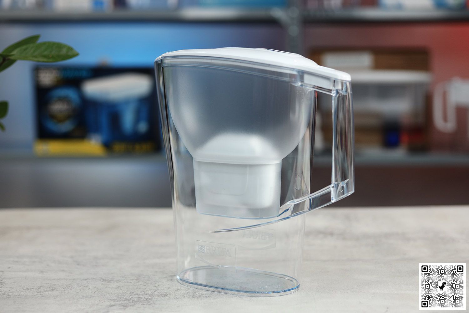 The Brita Aluna on a table, houseplant leaves on the left, water pictures on a shelf in background