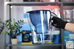 Gloved hand picking up the blue Brita Marella XL at its handle, houseplants and a shelf in the background