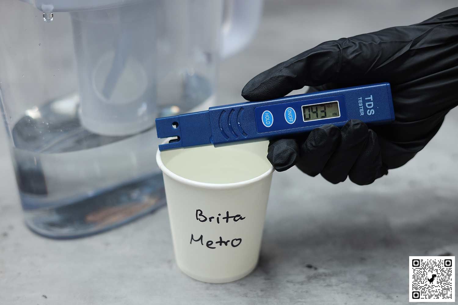 Gloved hand holding a water TDS meter over a paper cup labeled Brita Metro, the lower part of a water filter pitcher in the background