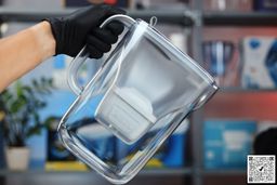 Gloved hand picking up the blue Brita Style by its handle, houseplants and a shelf in the background