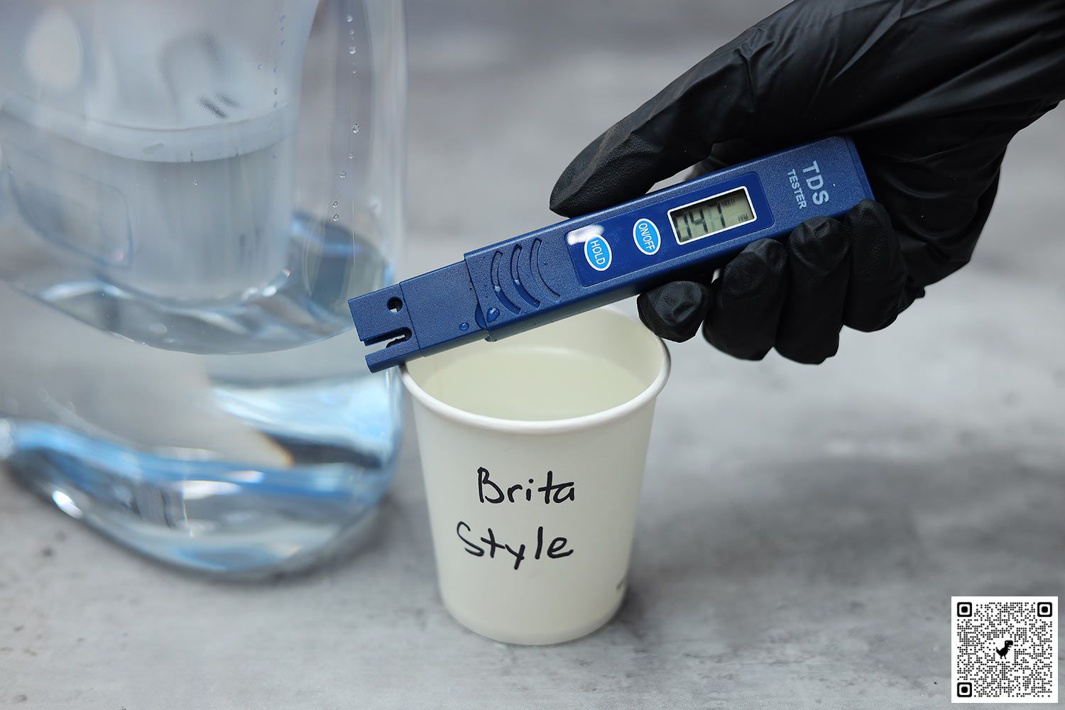 Gloved hand holding a water TDS meter over a paper cup labeled Brita Style, next to the lower part of a water filter pitcher