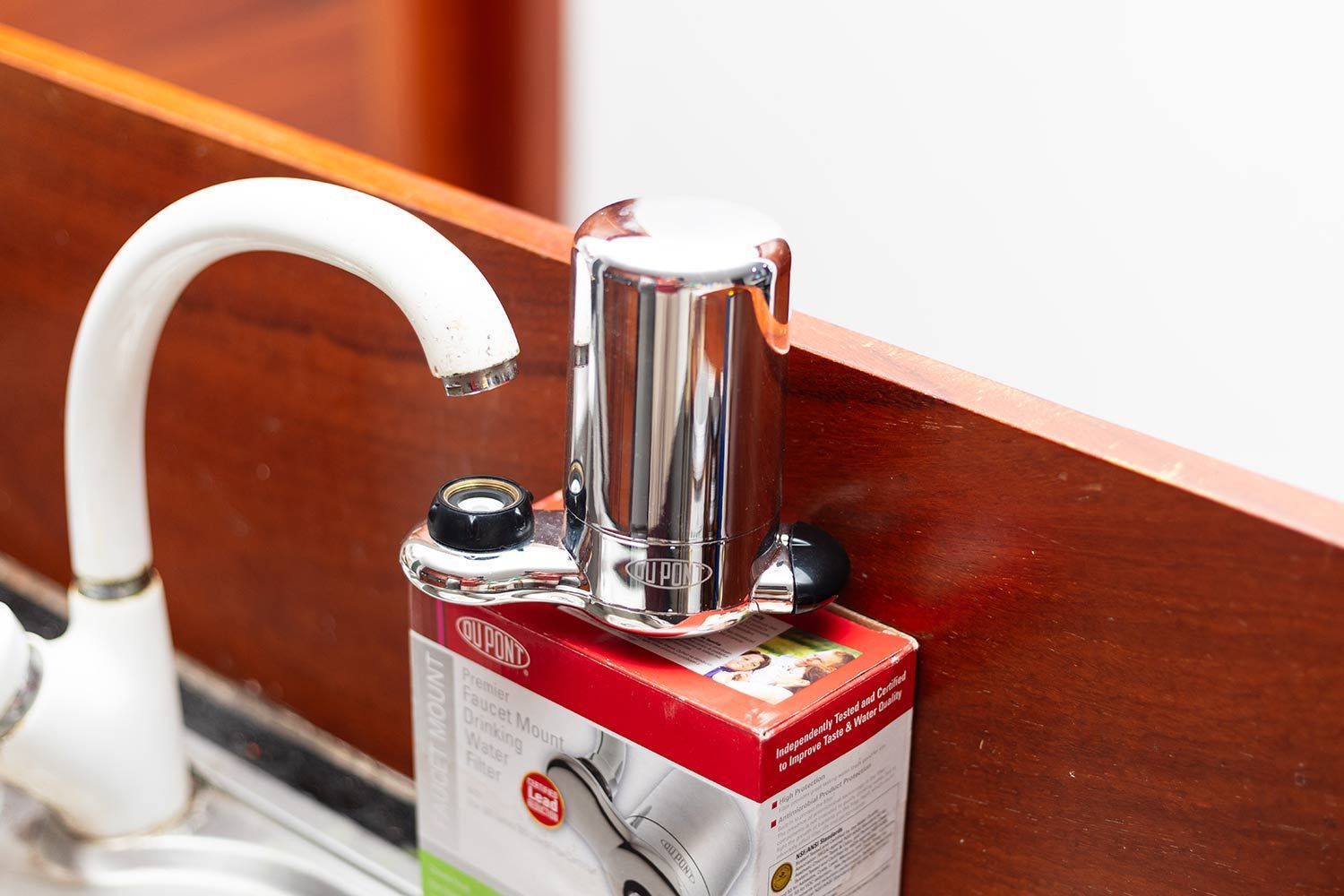 The DuPont faucet mount water filter sitting on top of its shipping box, below a white water faucet.