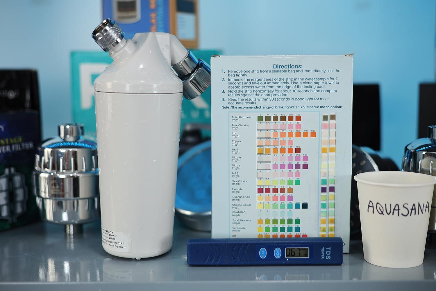 Water test kit with two used strips next to the Aquasana AQ-4100 shower filter, paper cup labeled Aquasana, and TDS tester