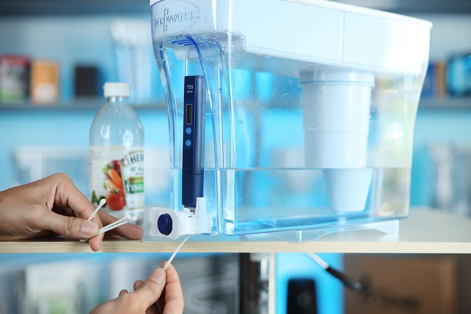 the ZeroWater 23 cup dispenser on a table, two hands holding Qtips to clean its spigot, a vinegar bottle in the background